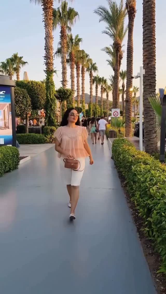 Woman wearing a peach blouse and white trousers walking along a palm-lined path.