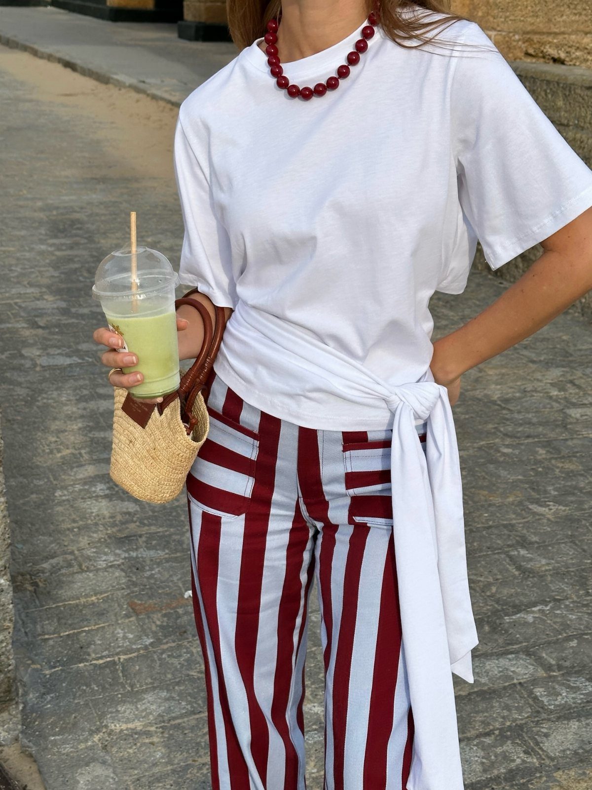 Person in white t-shirt and striped pants holding a green drink and straw bag.