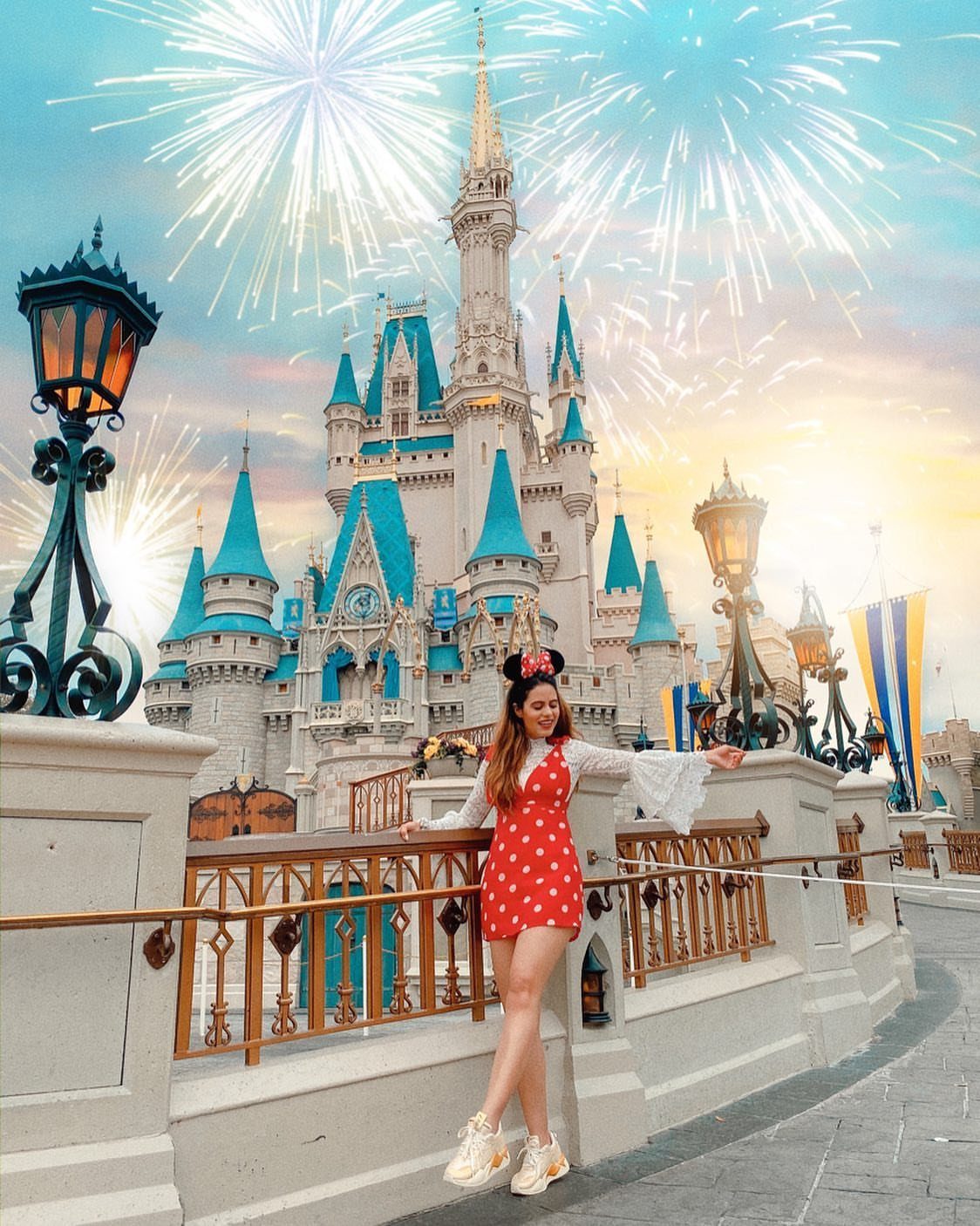 Woman in red polka-dot dress and white sneakers posing before a fairytale castle.