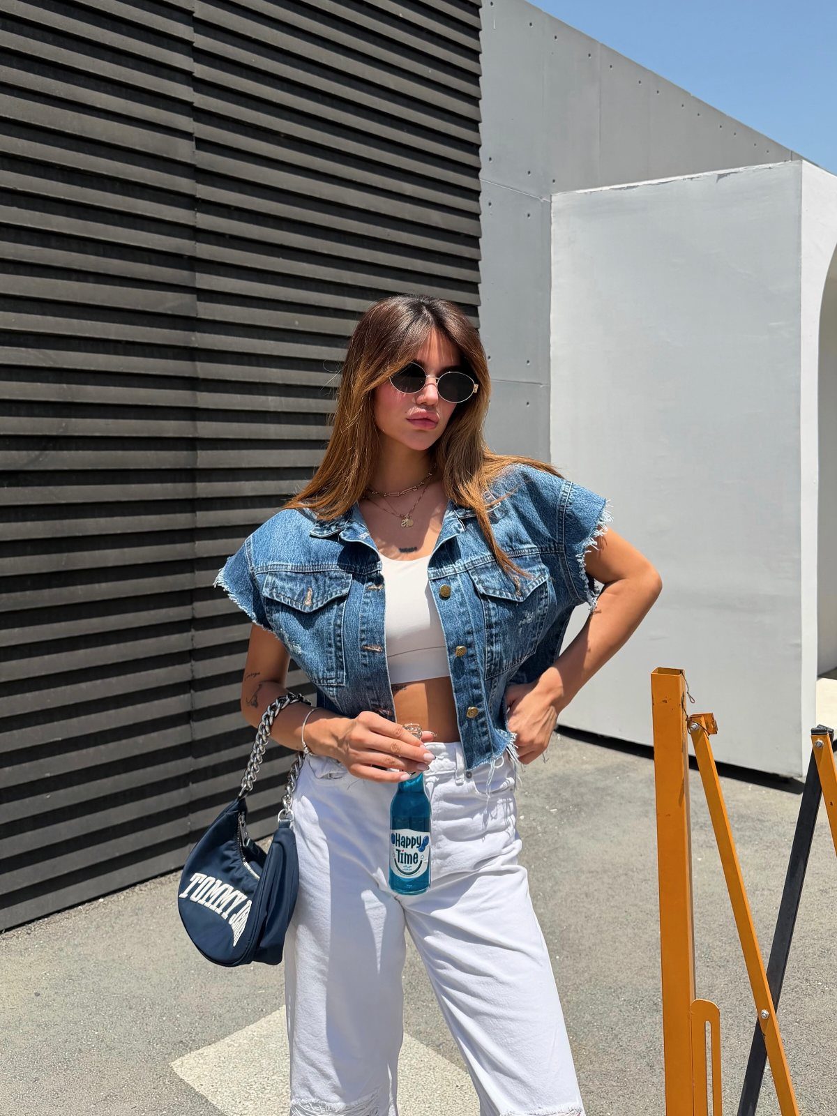 Woman in denim vest and white wide-leg trousers posing against a grey wall.