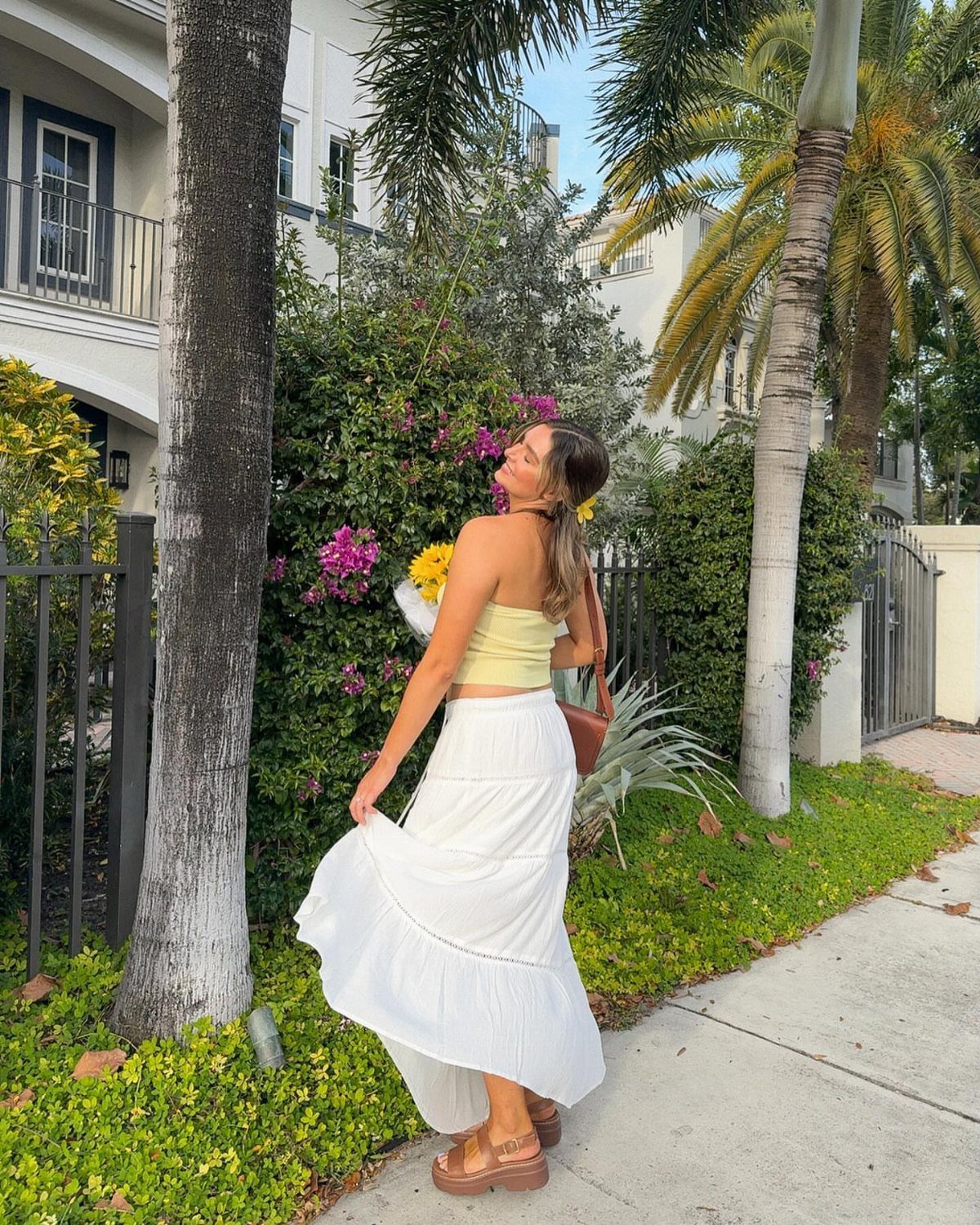 Woman in yellow tube top and white maxi skirt posing by palm trees.