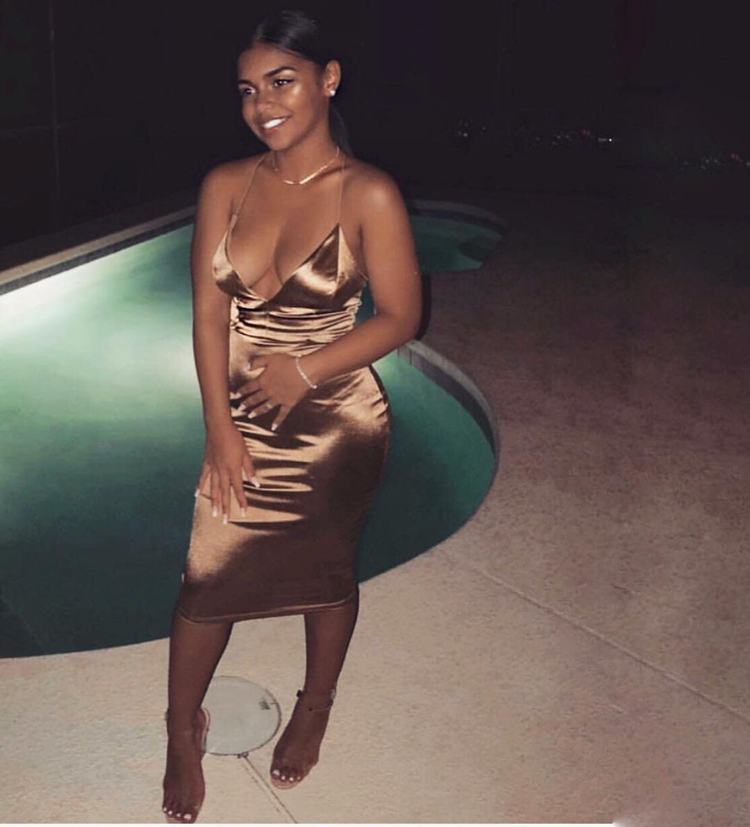A woman smiling while wearing a gold satin midi dress standing near a pool at night.