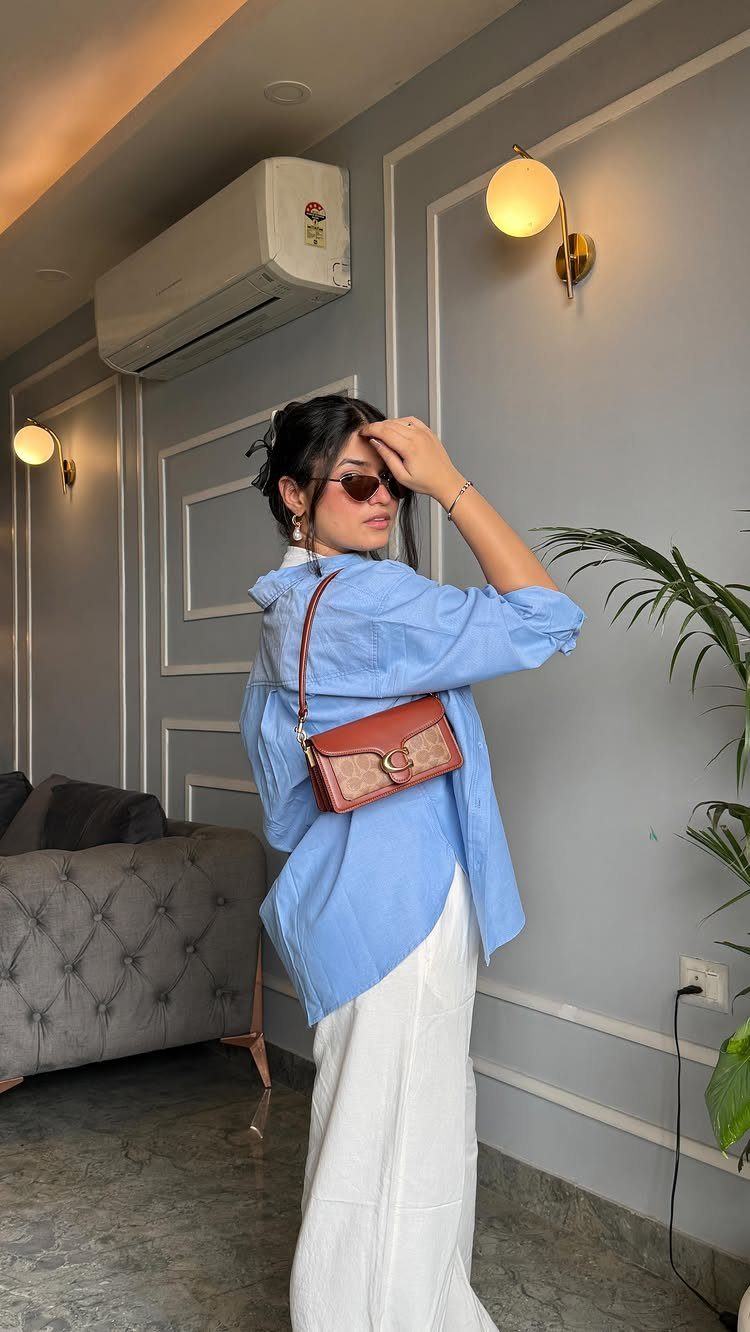Woman wearing a blue button-down shirt and white linen pants with a brown shoulder bag.