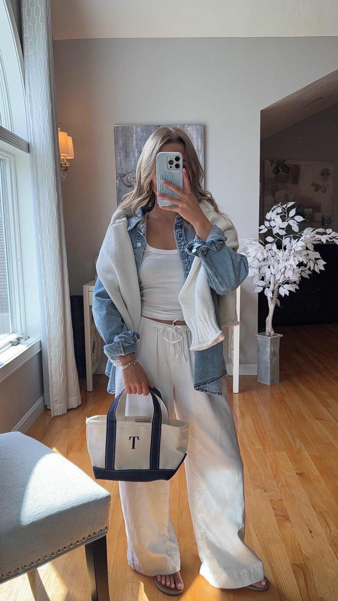 Woman posing in a mirror wearing white linen pants, a white tank top, and a denim jacket.