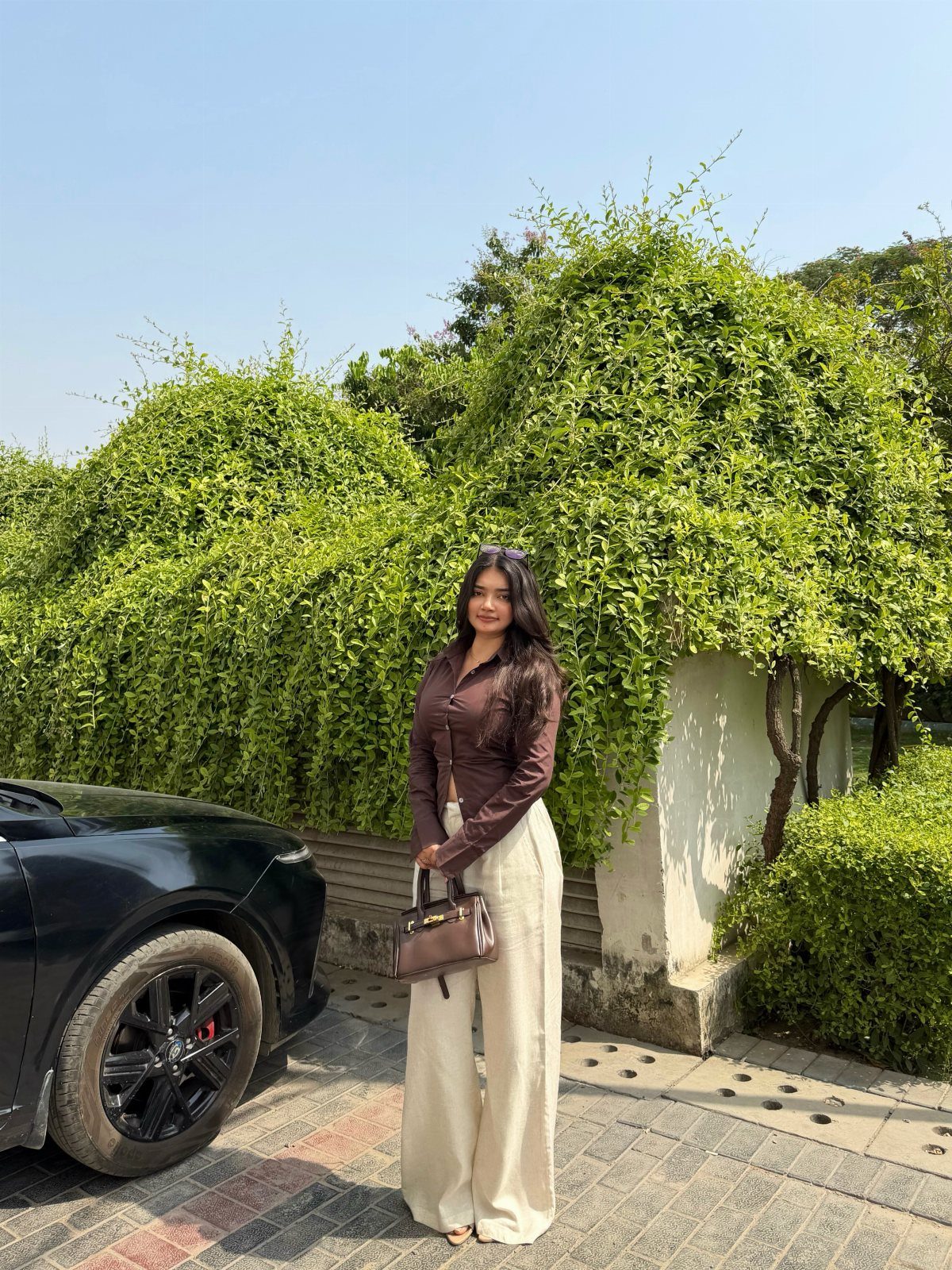 Woman wearing cream linen pants and a fitted brown button-down shirt standing by green bushes.