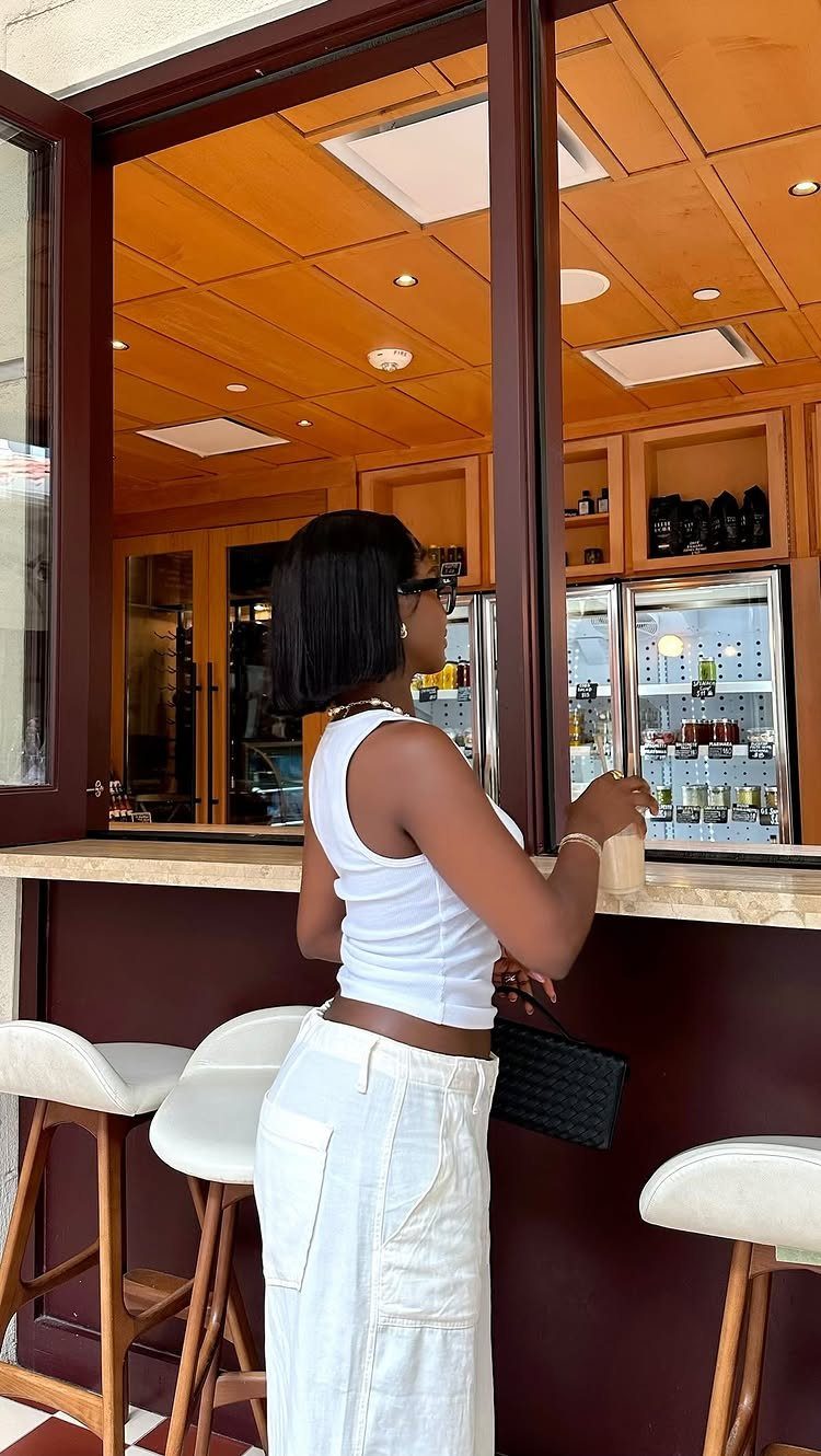 Woman in a white tank top and white linen pants standing at a bar.
