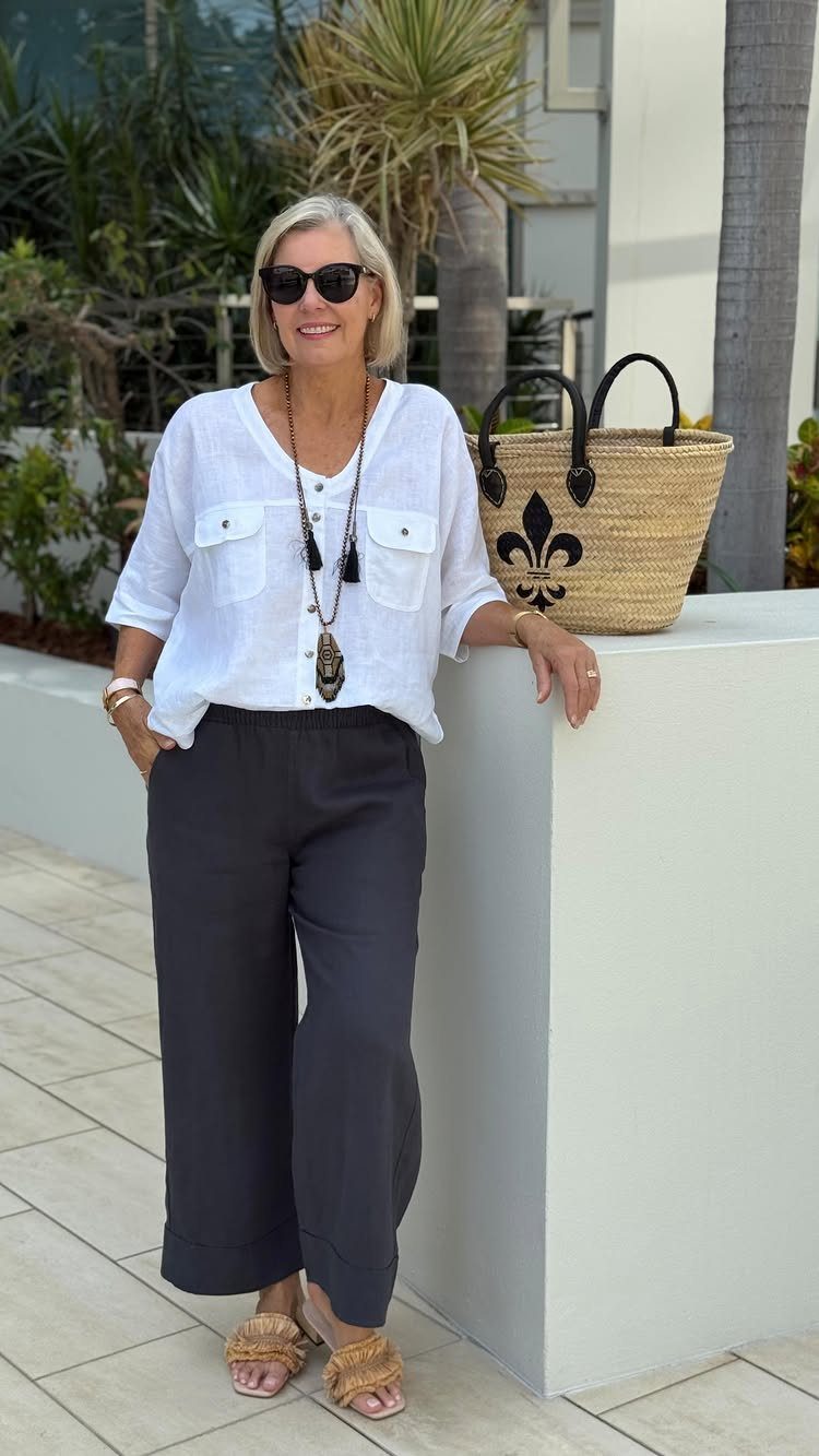 Woman in white linen shirt and black wide-leg pants standing next to a straw tote bag.