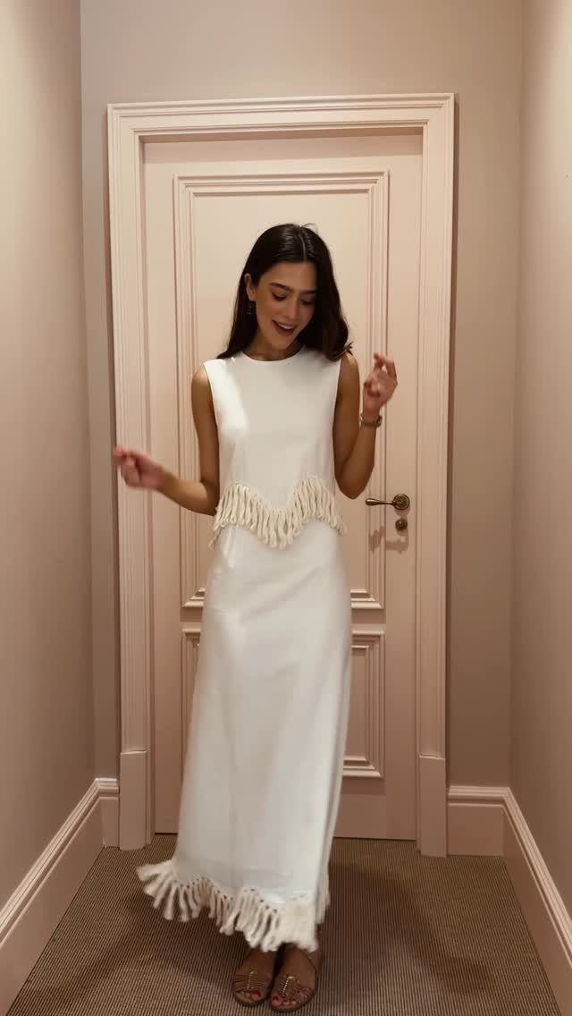 A woman posing in a long, sleeveless white linen dress with fringe trim.
