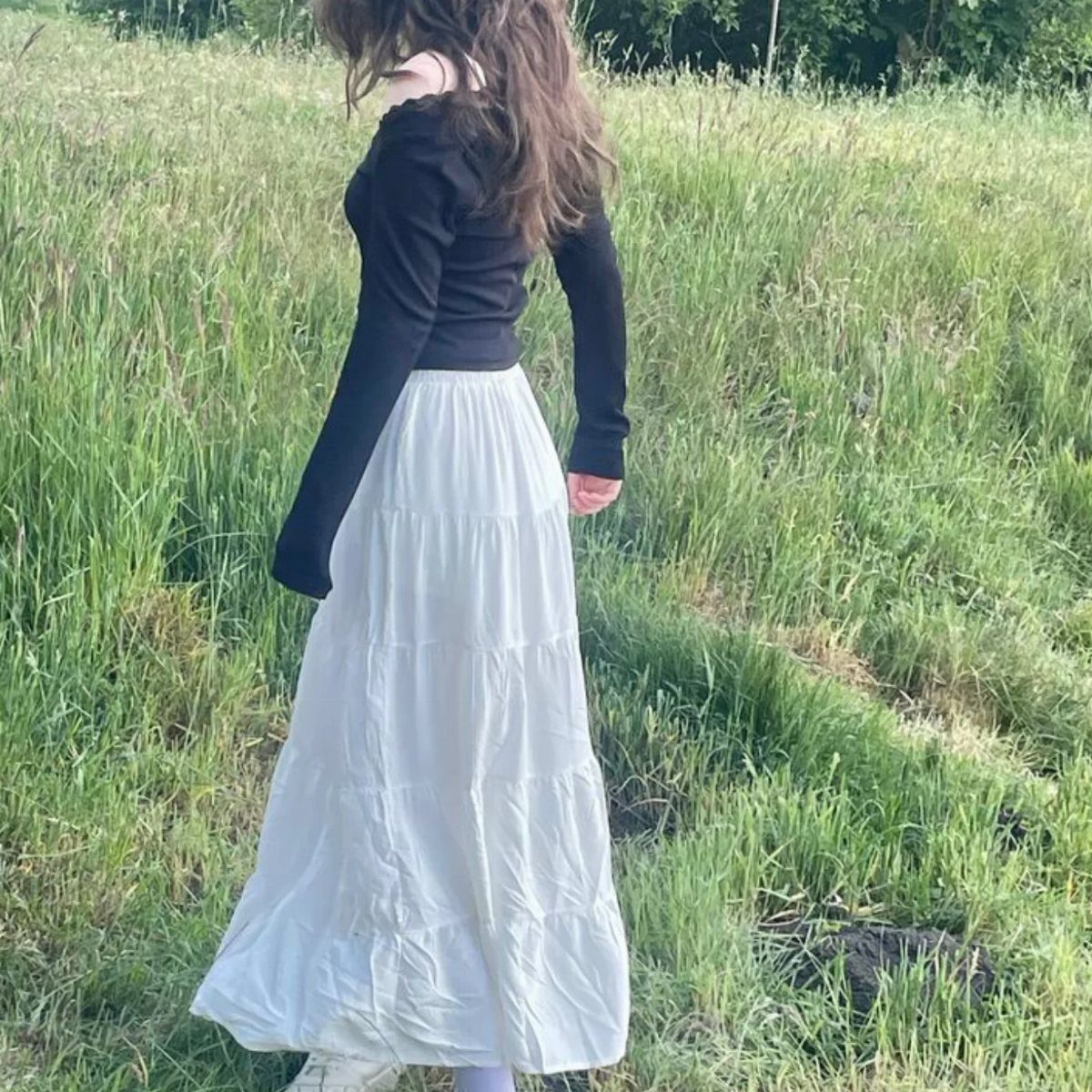 Woman in a grassy field wearing a black long-sleeved top and white maxi skirt
