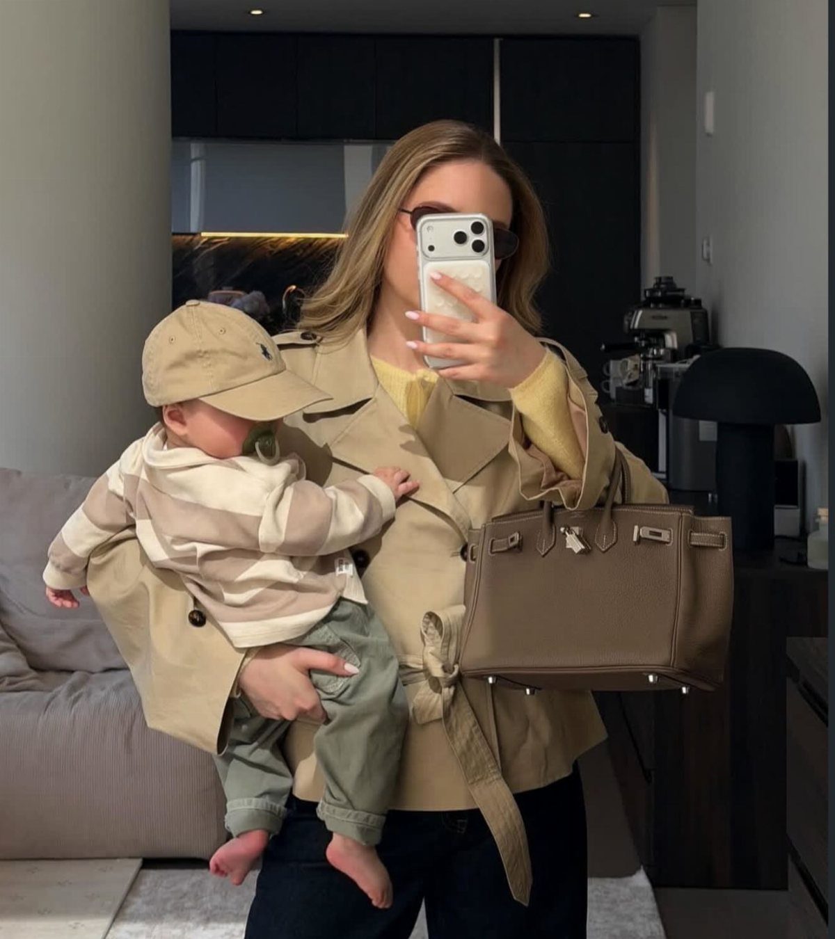 10 Outfit Ideas for Mom: From Classic Days to Mom's Nights Out 4 Woman in a tan belted trench coat holding a baby and a large leather handbag.