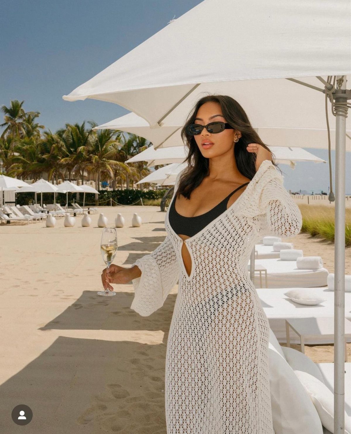 Outfit for Vacation: 13 Ideas for Every Island Moment 11 Woman in a white crochet cover-up holding champagne on a sunny beach with umbrellas.