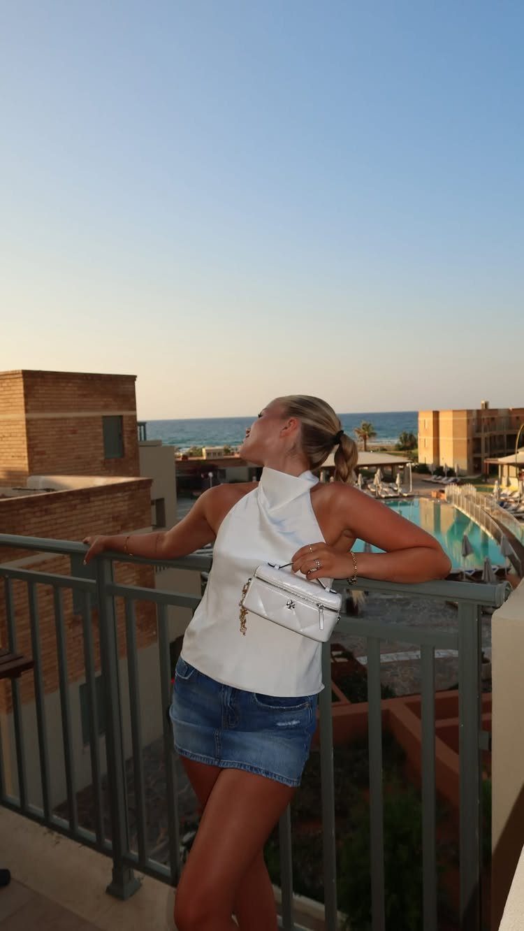 Outfit for Vacation: 13 Ideas for Every Island Moment 13 Woman in a white halter top and denim skirt leaning on a balcony railing overlooking the coast.