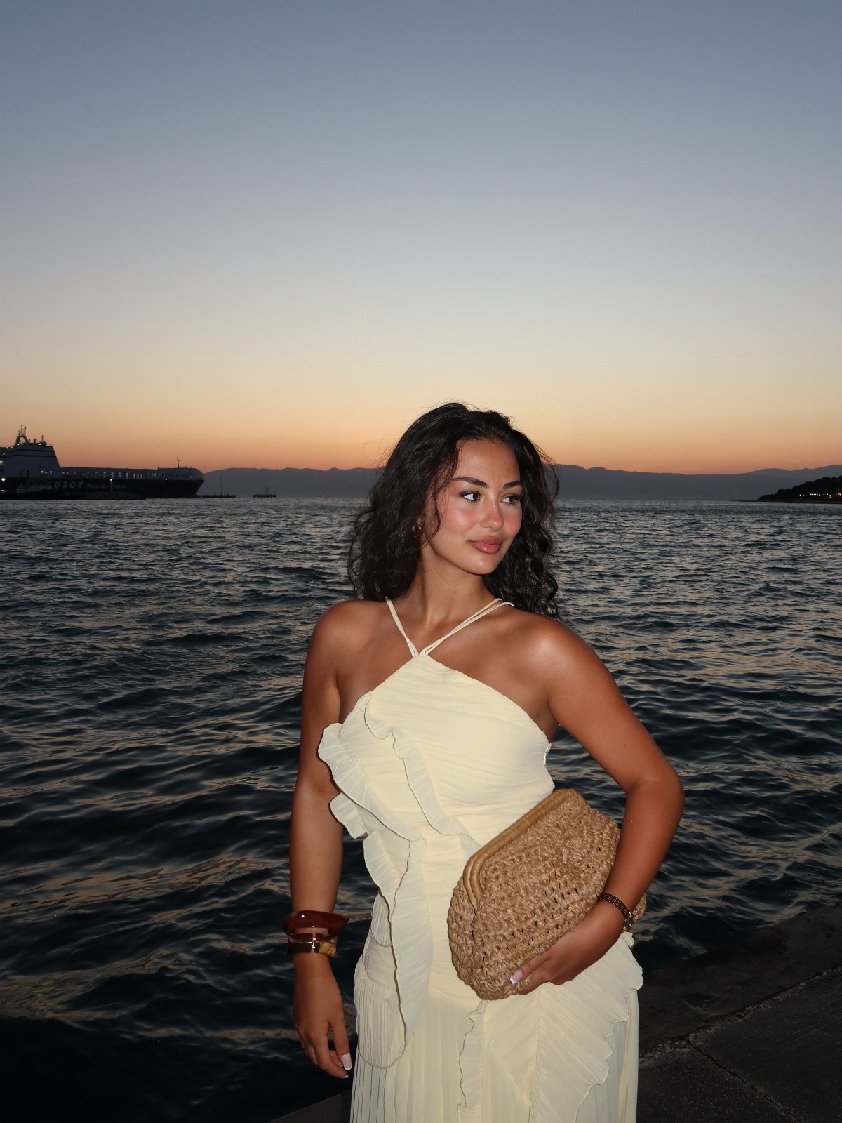 Outfit for Vacation: 13 Ideas for Every Island Moment 1 Woman in a cream ruffled halter dress posing by the ocean during sunset.