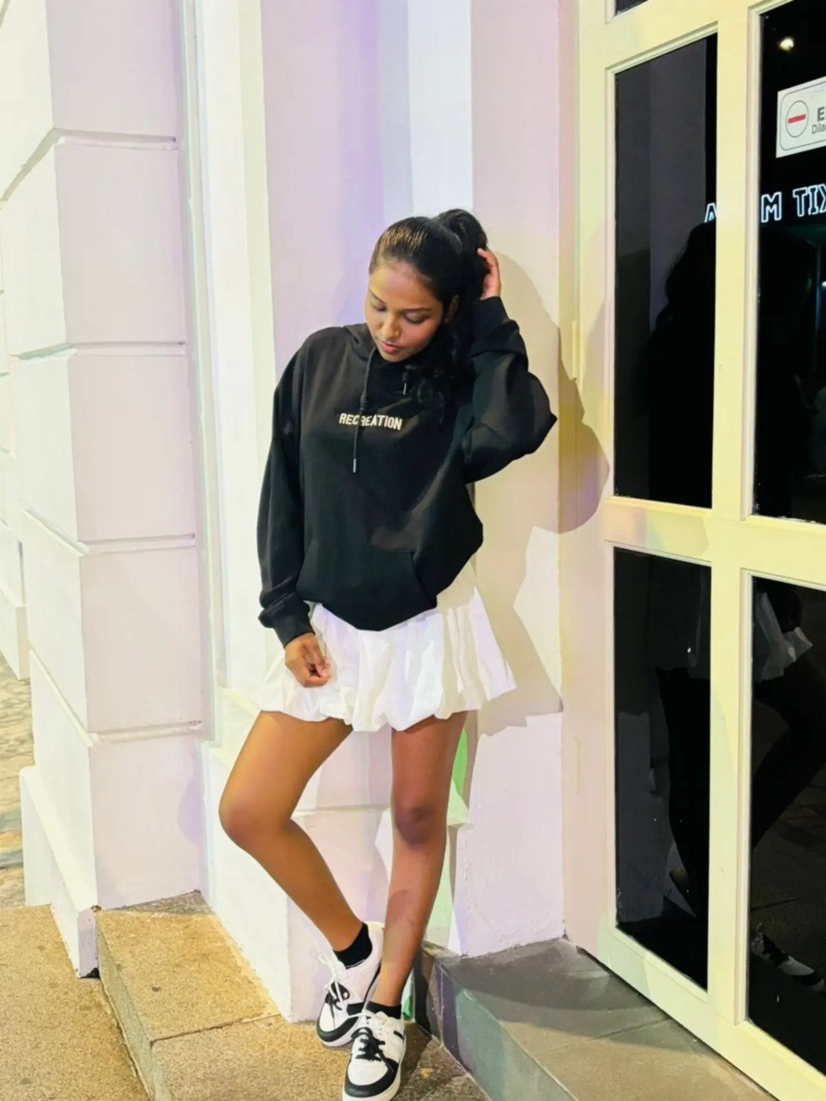 Woman in black oversized hoodie and white pleated mini skirt posing by white wall.