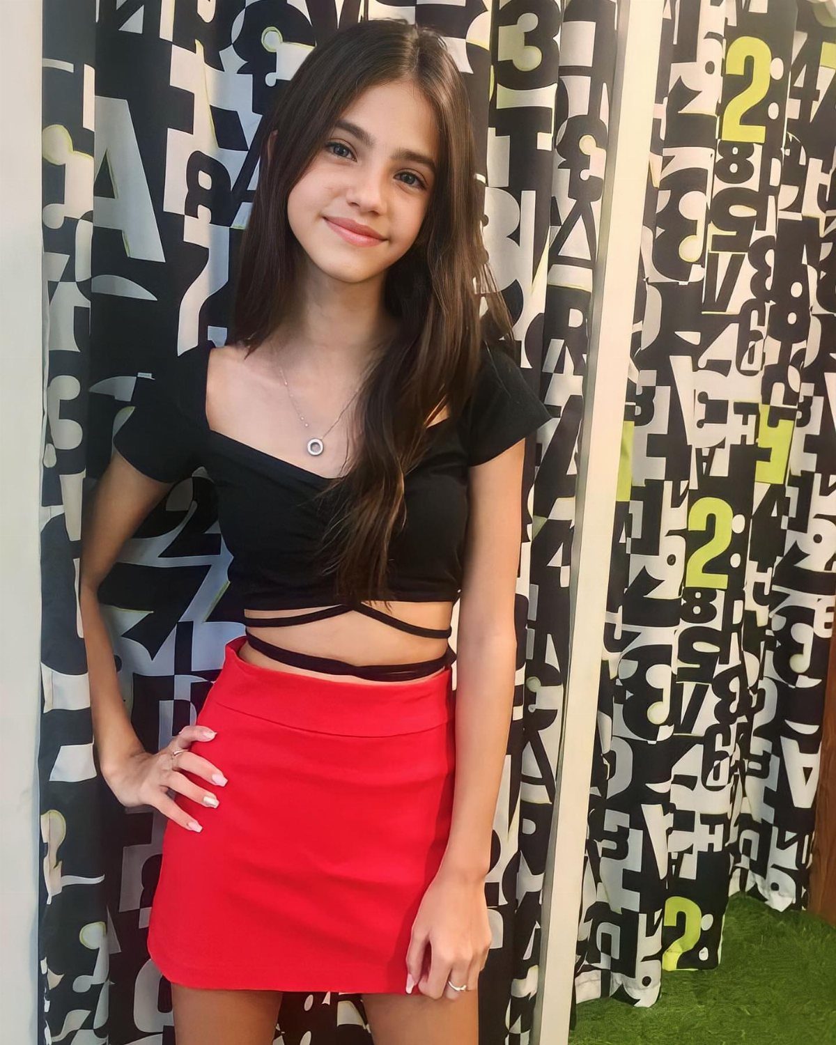 11 Outfits for Teens: Street to Semiformal Style Ideas 9 A smiling girl wearing a black crop top and a red mini skirt standing before patterned curtains.