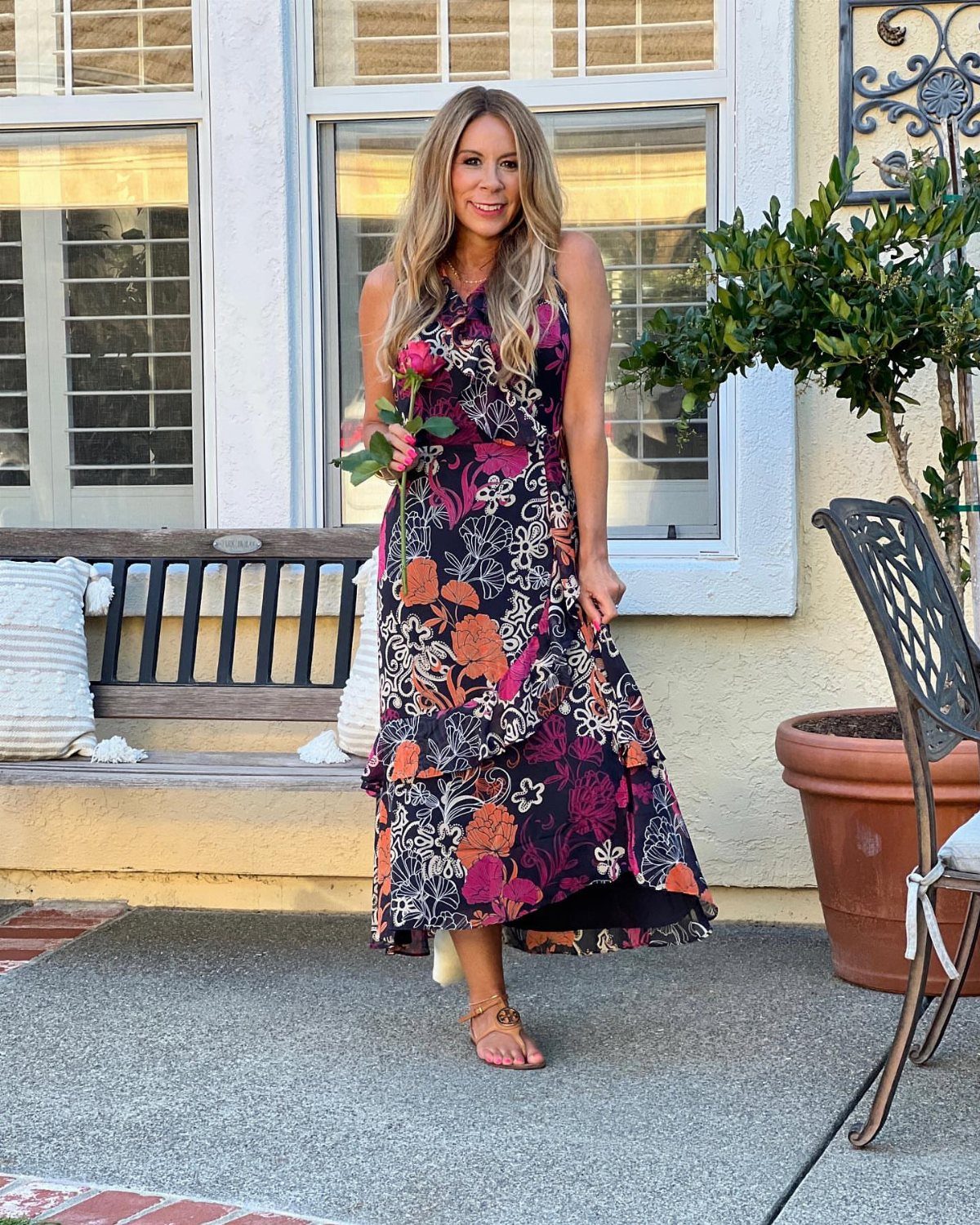 Woman in a dark floral sleeveless maxi dress standing on a patio holding a rose.