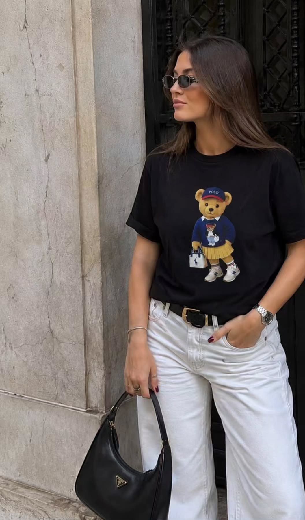 Woman in black bear graphic t-shirt and white wide-leg trousers holding a black handbag.