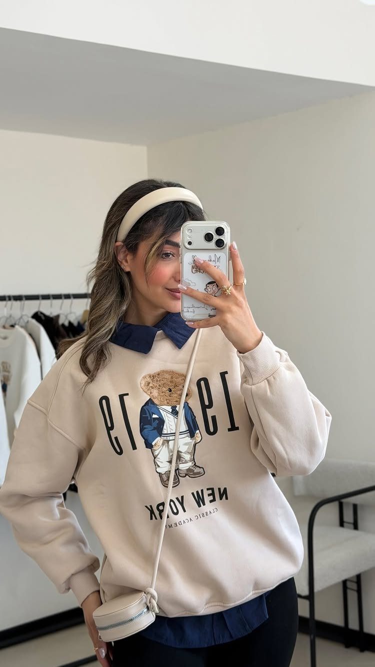 Woman wearing a beige bear graphic sweatshirt layered over a blue collared shirt.