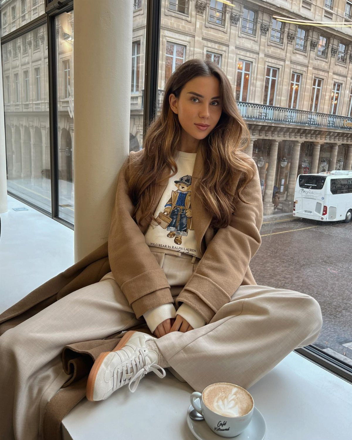 Woman wearing a bear graphic sweater and beige wide-leg trousers sitting by a window.