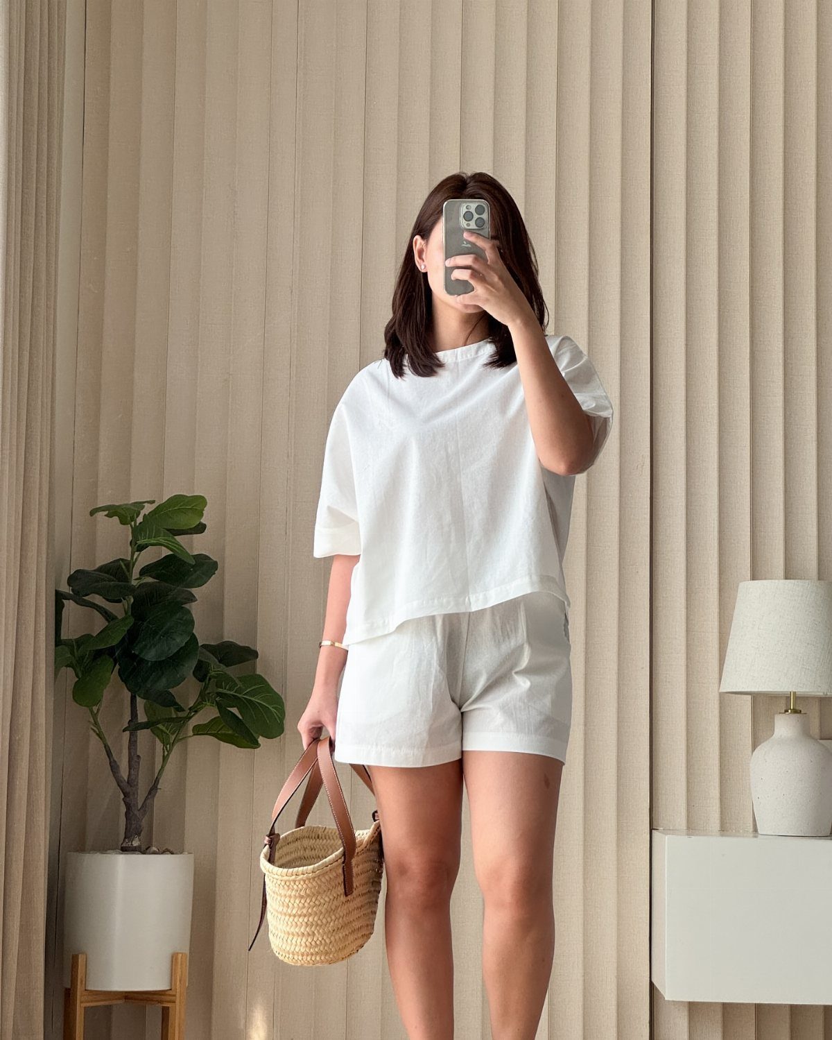 15 Preppy Summer Outfits to Wear All Season Long 7 Woman wearing a white linen shirt and matching shorts with a straw tote bag.