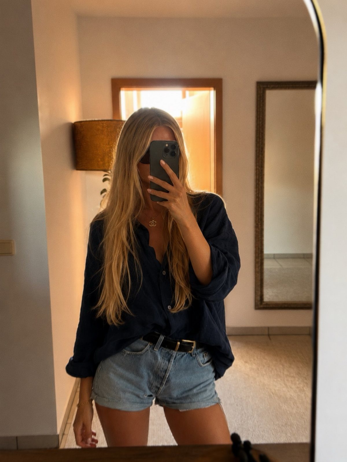 15 Preppy Summer Outfits to Wear All Season Long 4 Woman wearing a navy blue button-down shirt and light-wash denim shorts in a mirror selfie.