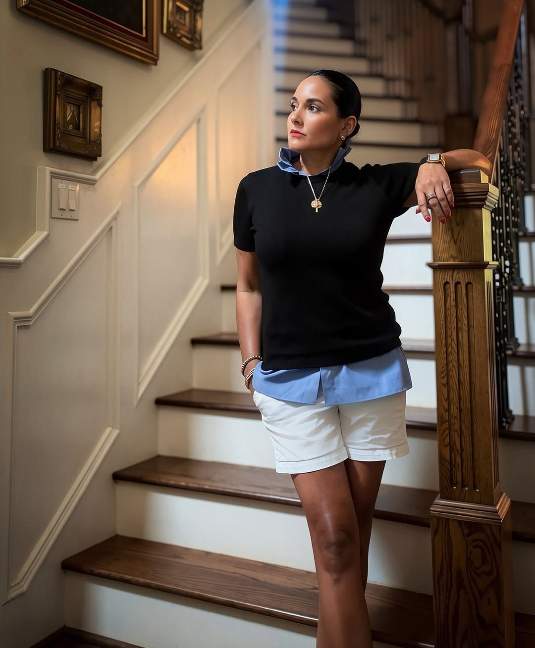 15 Preppy Summer Outfits to Wear All Season Long 9 Woman wearing a black sweater, light blue shirt, and white tailored shorts on stairs.