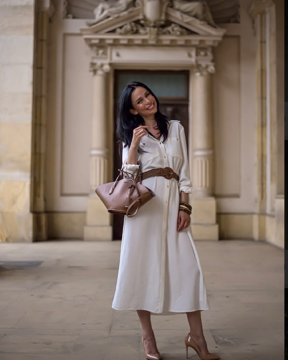 Woman in a white belted shirt dress and nude heels before a stone building.