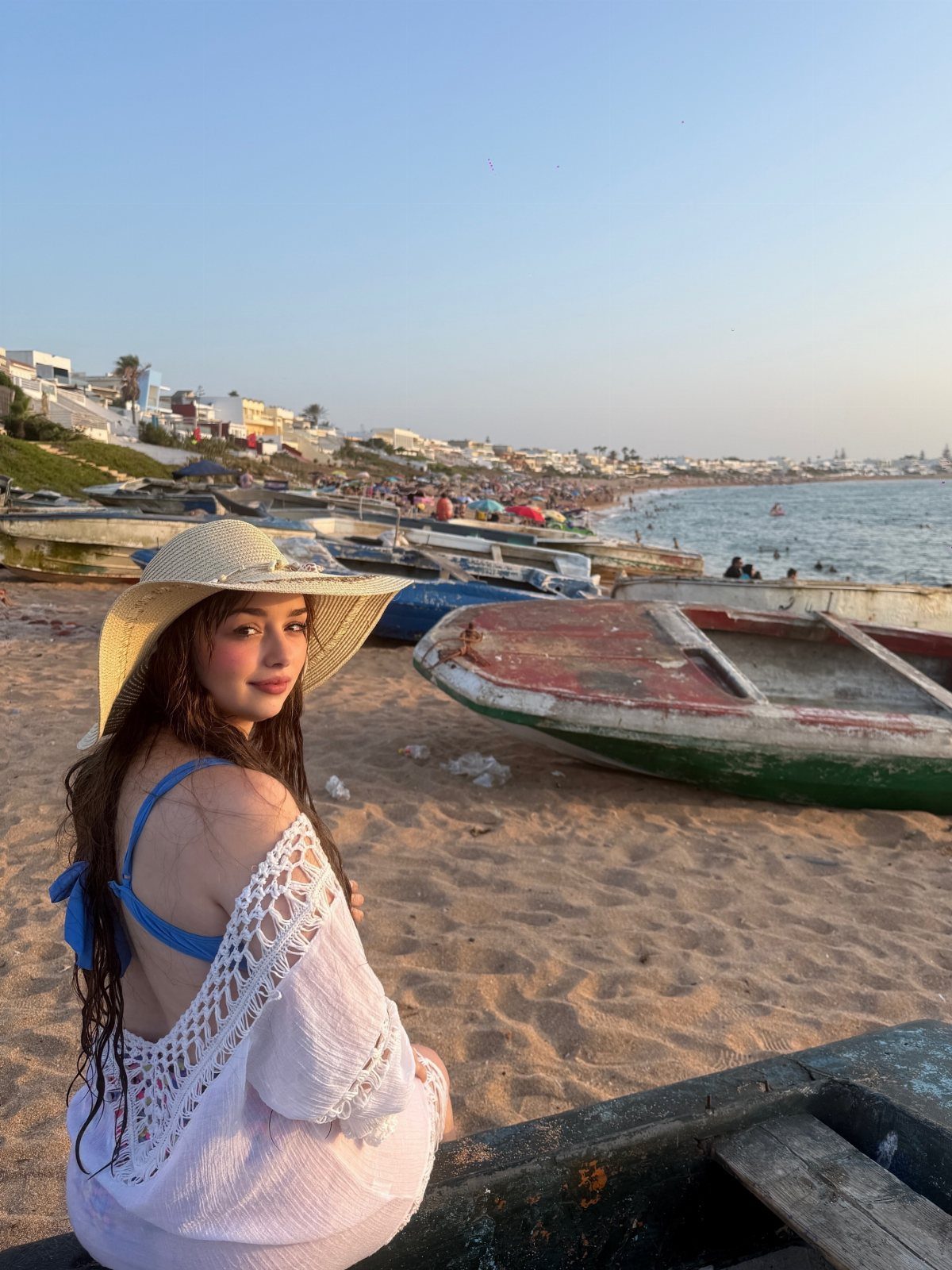 Woman in white crochet beach cover-up and straw hat posing by beach boats.