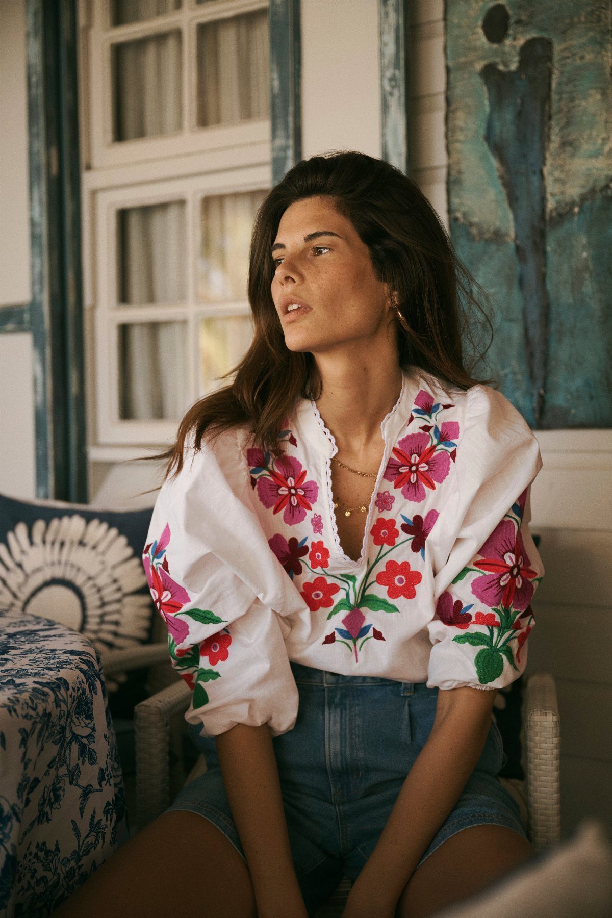 Woman wearing a white floral embroidered blouse and blue denim shorts on a porch.
