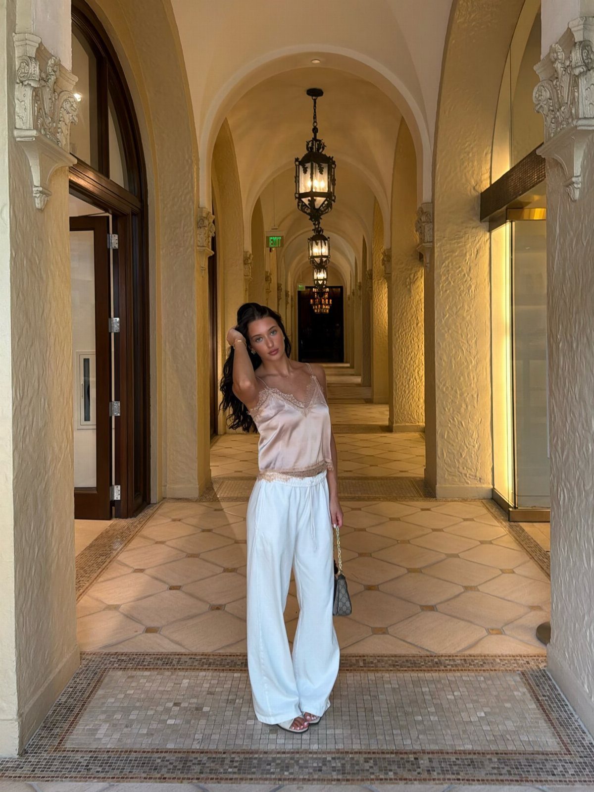 Woman wearing a champagne satin camisole and white wide-leg trousers in a vaulted hallway.