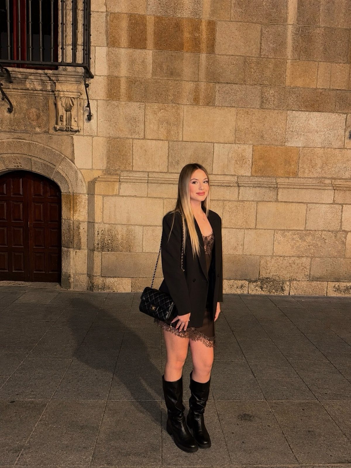 Woman wearing a black blazer over a black slip dress and knee-high boots.