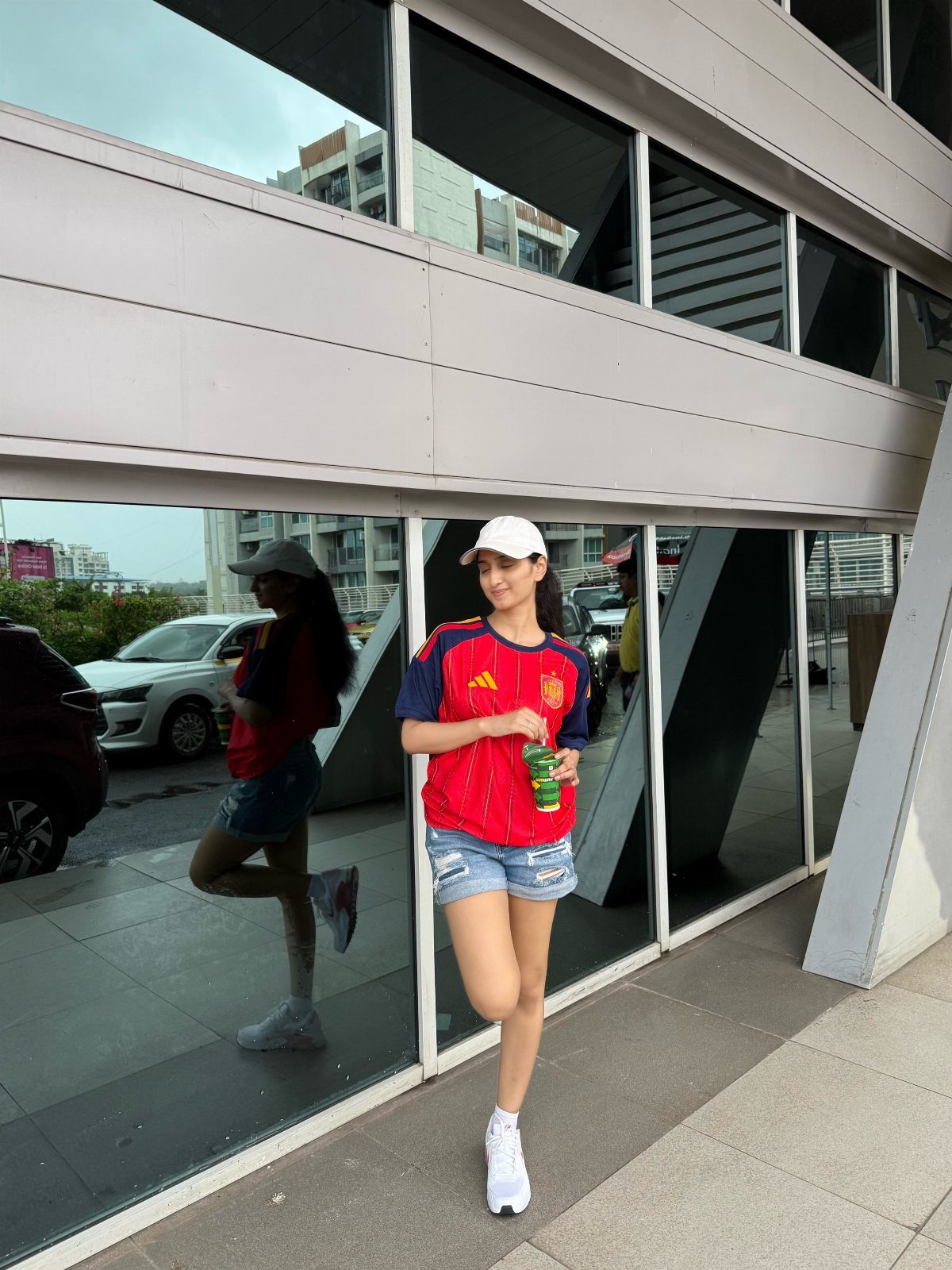 Woman wearing a red Spain football jersey and denim shorts posing by a mirror.