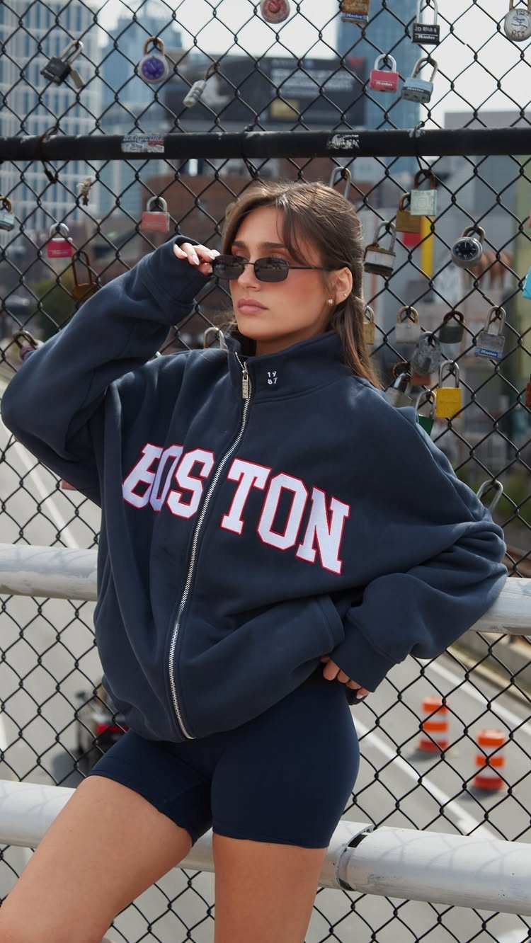 12 Sporty Chic Outfits That Work Beyond the Gym 1 Woman in navy Boston zip-up hoodie and matching biker shorts posing by a fence.
