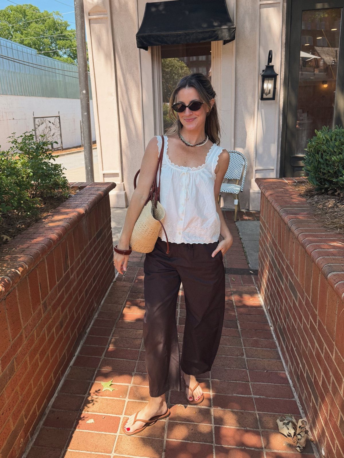11 Spring Summer Outfits You'll Reach for All Season Long 8 Woman wearing a white eyelet top and dark brown wide-leg trousers on brick path.