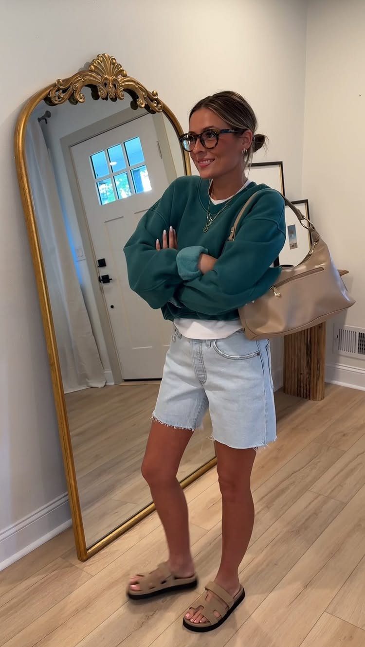 11 Spring Summer Outfits You'll Reach for All Season Long 4 Woman wearing a teal sweatshirt, white t-shirt, denim shorts, and brown sandals.