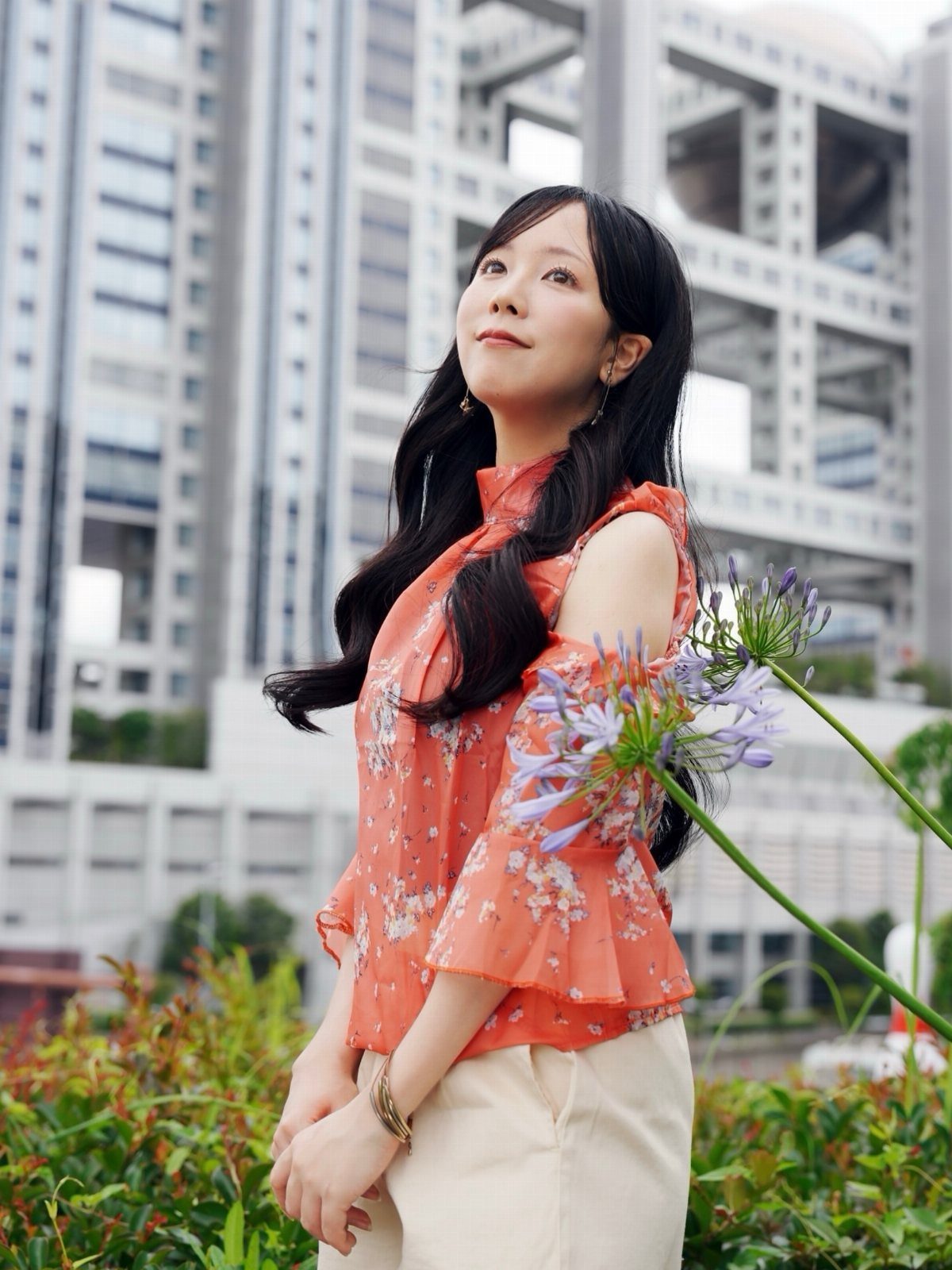 Woman wearing an orange floral blouse and cream pants standing before city buildings.