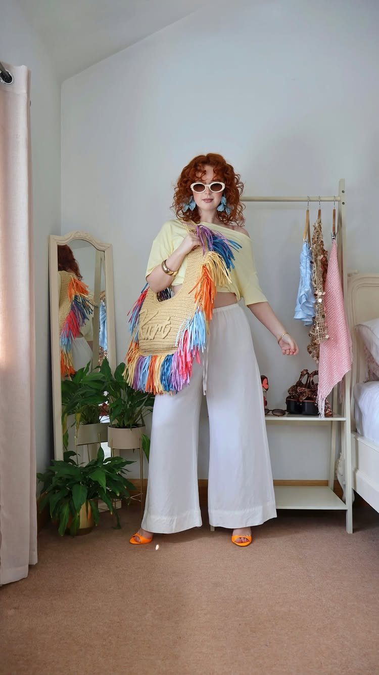 Woman wearing white wide-leg linen pants, yellow top, and colorful fringed straw bag.