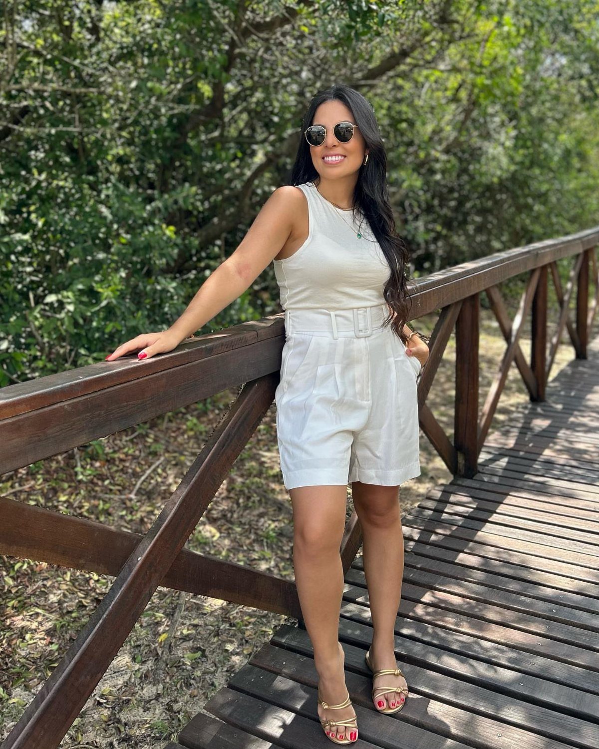 Woman in white sleeveless top and belted shorts posing on a wooden bridge.