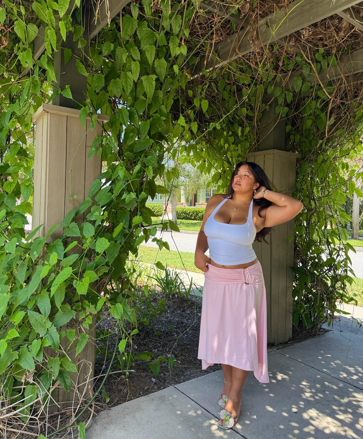 Woman wearing a white cropped tank top and pink midi skirt under a pergola.