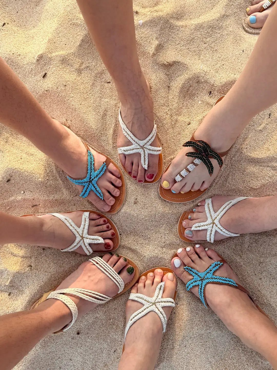 Multiple people wearing starfish-themed beaded sandals arranged in a circle on the sand.