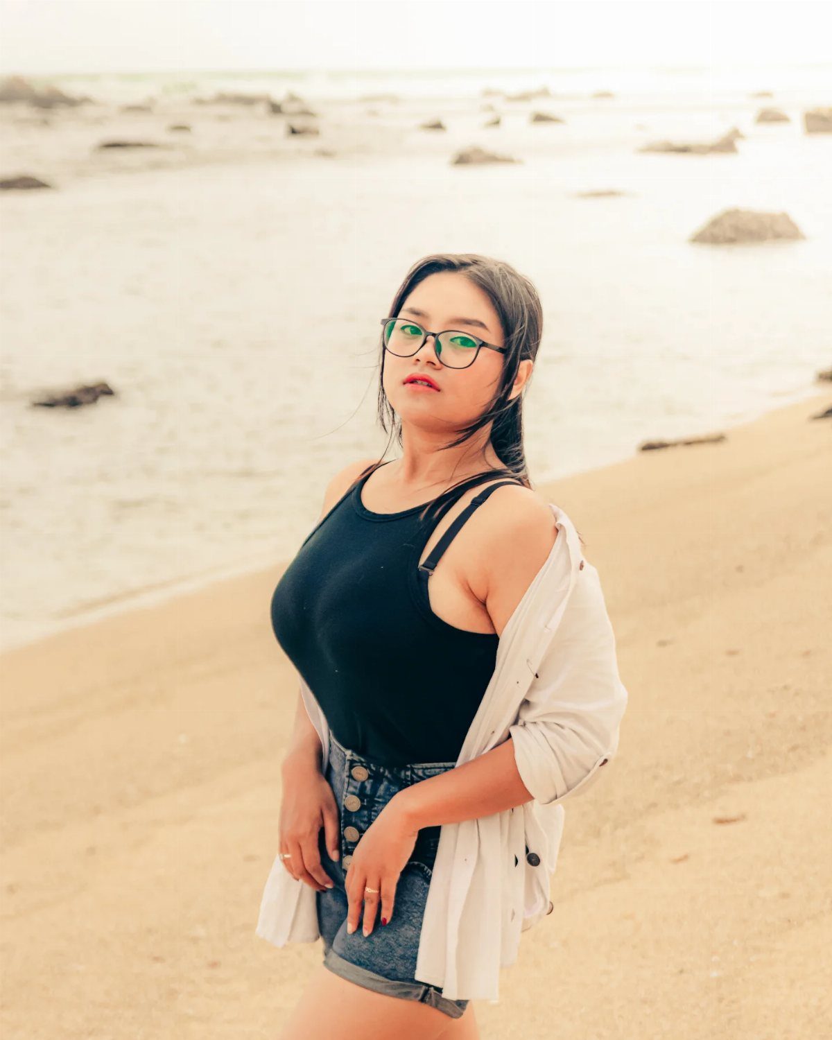 A woman in a black tank top and denim shorts posing on a sandy beach.