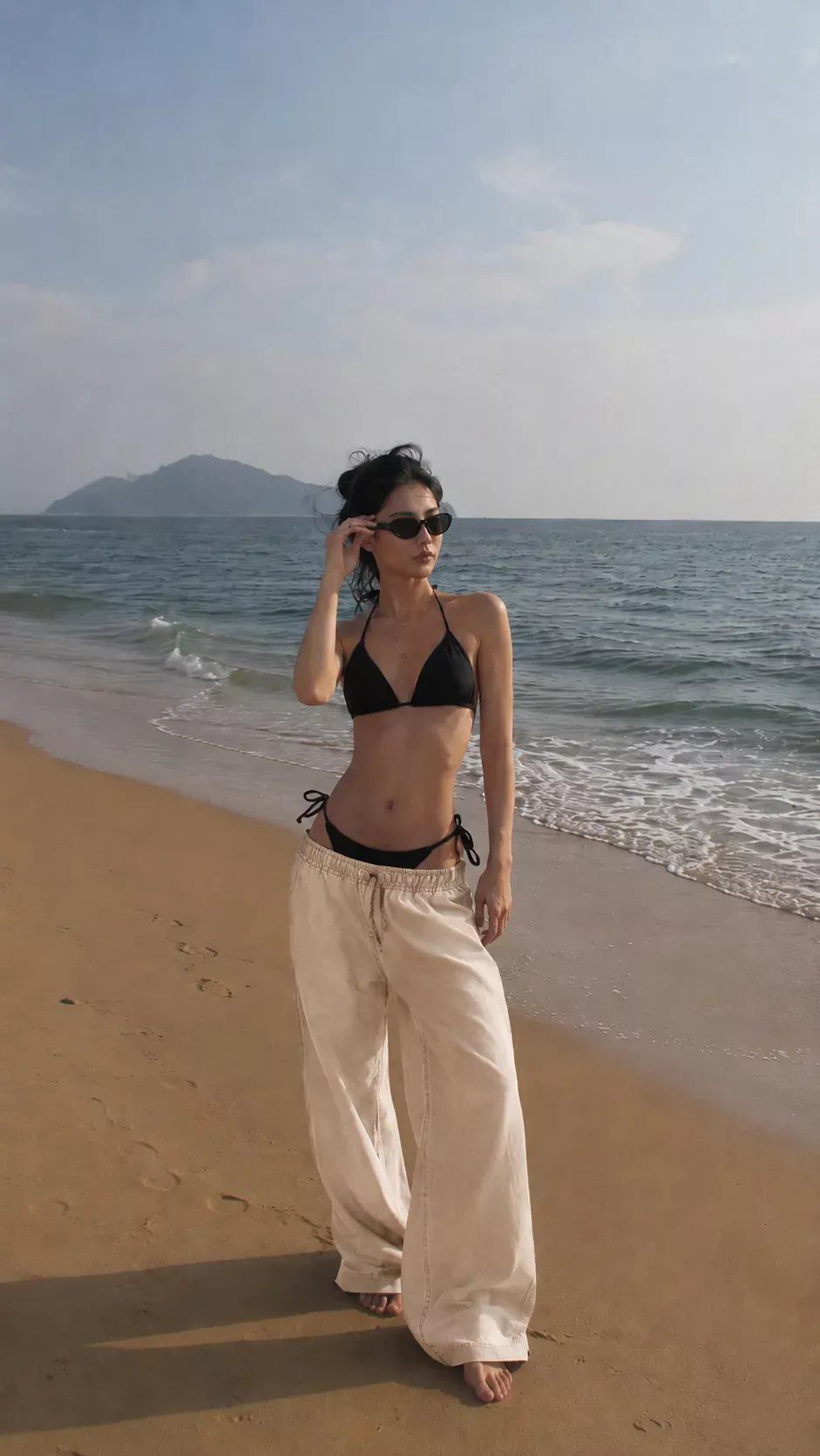 Woman in a black bikini and cream wide-leg trousers standing on a sandy beach.