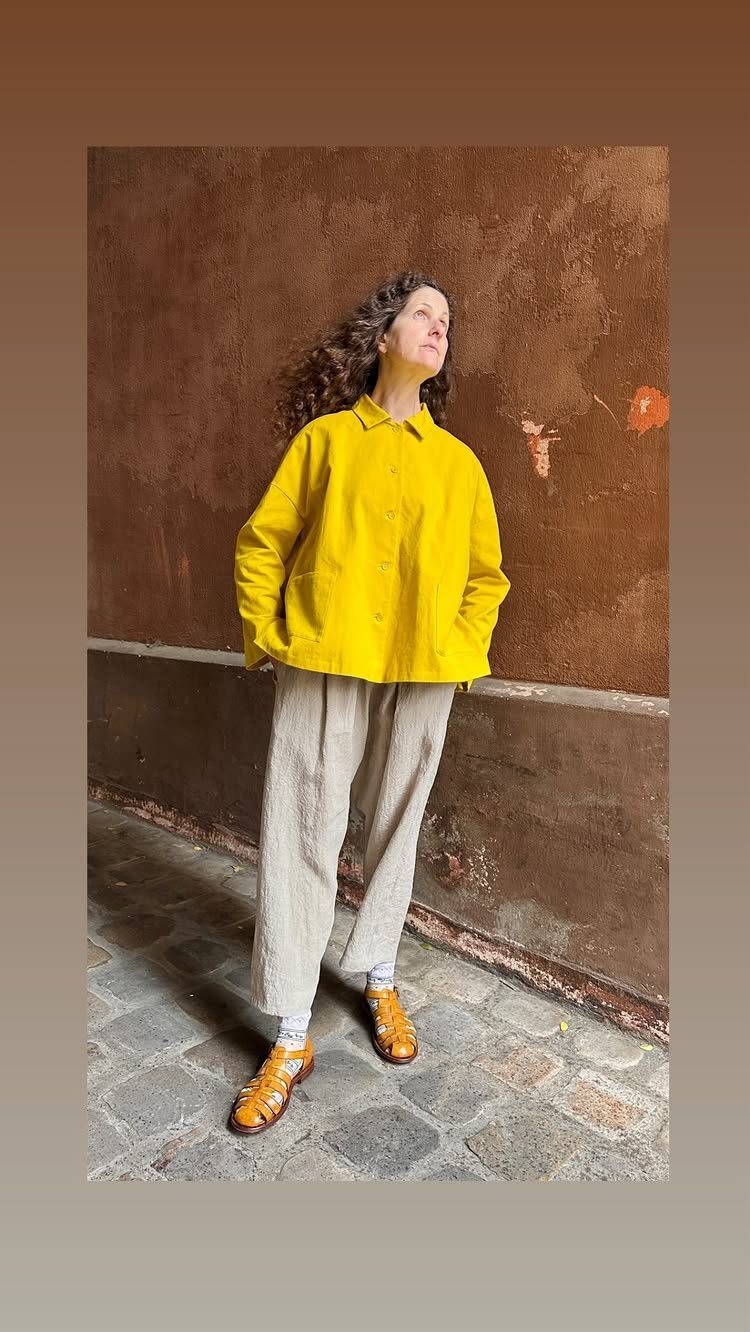 Woman wearing a bright yellow jacket and beige wide-leg trousers standing by a wall.