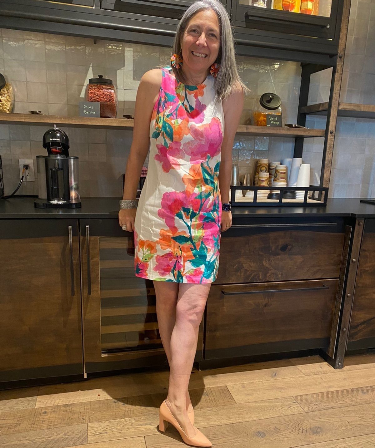 Woman with grey hair wearing a floral summer dress and tan heels.
