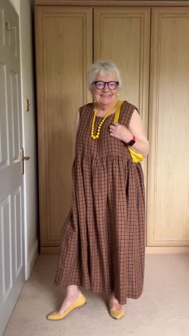 Older woman wearing a brown checkered maxi dress and yellow accessories standing indoors.