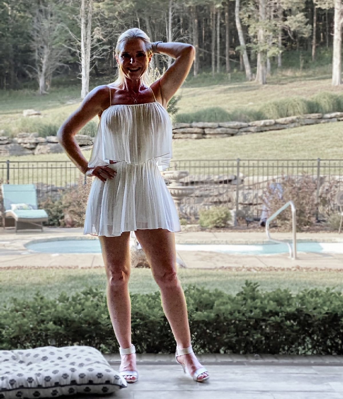Mature woman wearing a short white summer dress and white sandals by a pool.