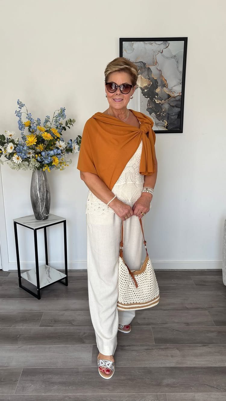 Woman over 60 wearing a white jumpsuit and orange wrap top with handbag.