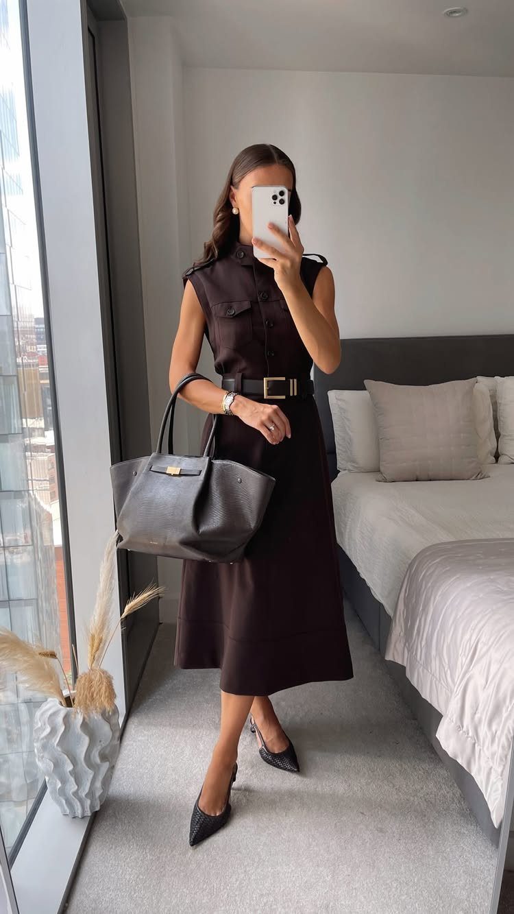 Woman in a dark brown sleeveless midi dress and black heels holding a large handbag.