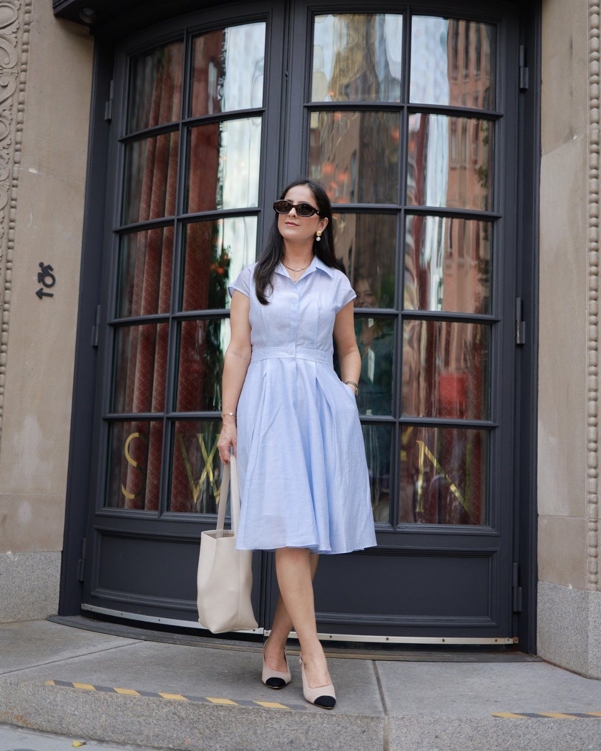 A woman posing in a light blue short-sleeved shirtdress and black heels in front of a glass door.