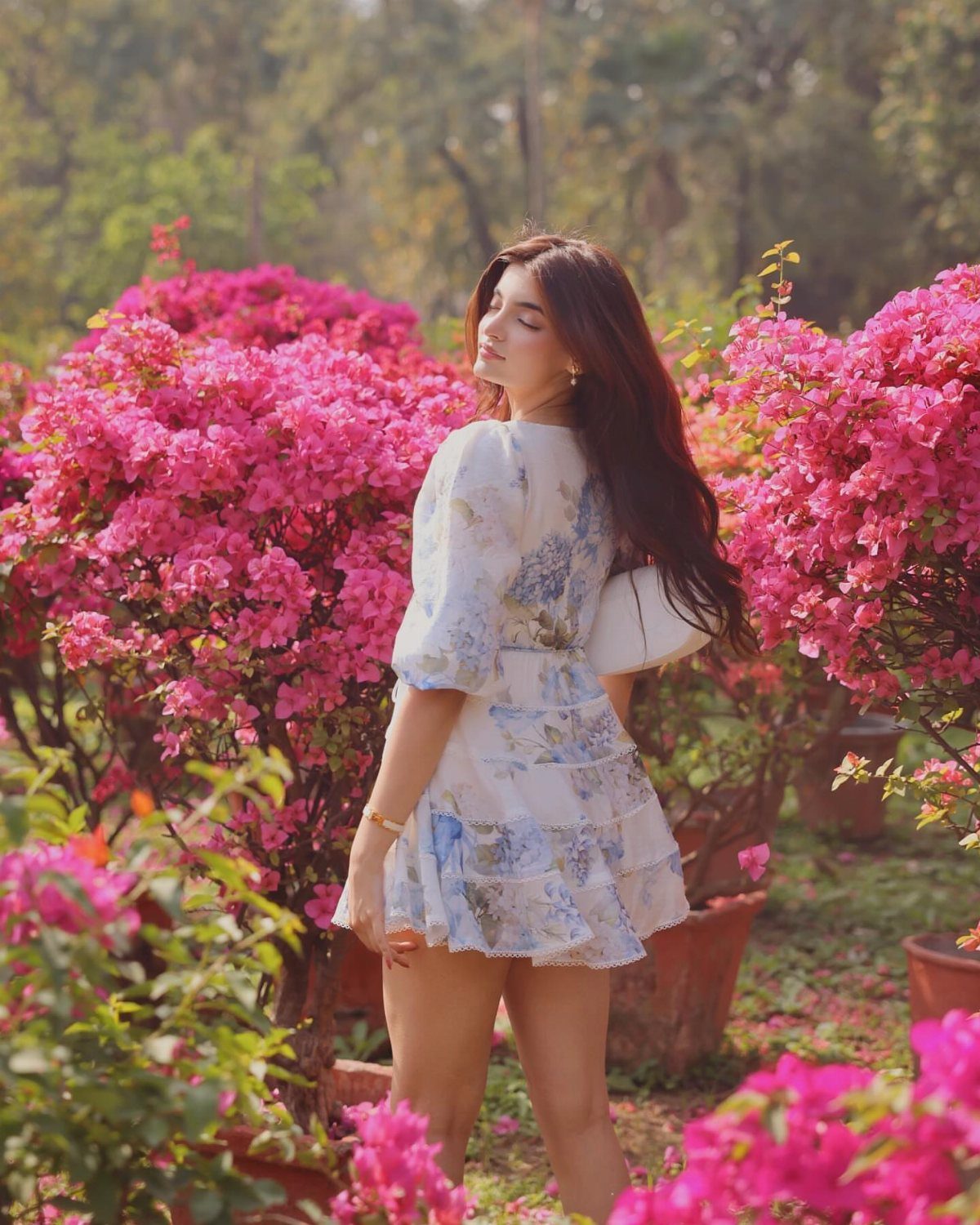 Woman in a white floral sundress standing amidst bright pink flowering bushes in a garden.